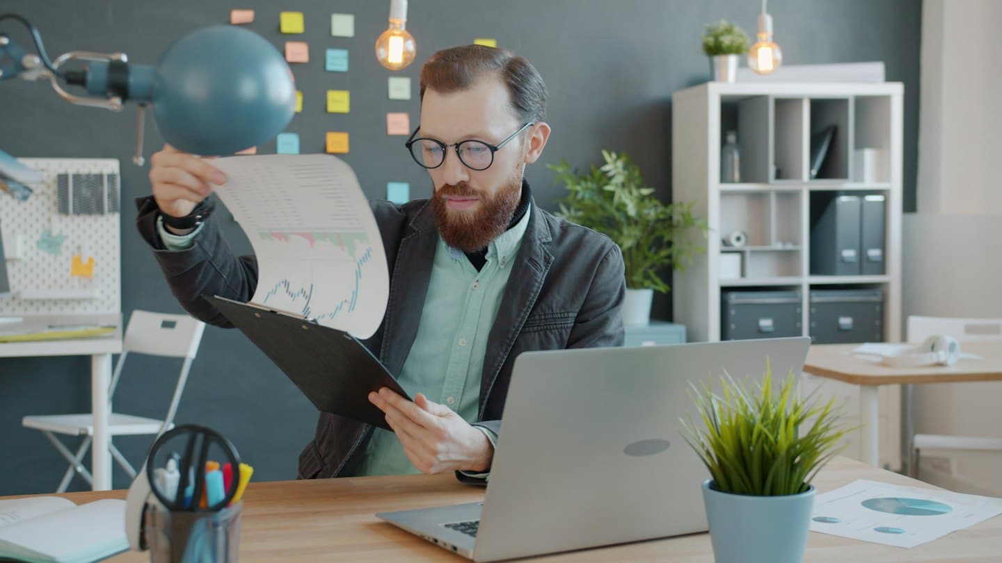 Man reviews financial graphs on paper at his wooden desk.